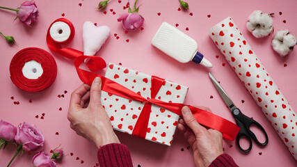 Obraz na płótnie Canvas Step 10.Step-by-step instructions for wrapping gifts for Valentine's Day. Woman wraps a gift with wrapping white paper in heart and seals the edges with glue on a pink background top view, flat lay