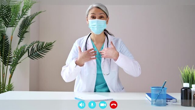 Webcam screen view. Mature gray-haired female doctor wears medical mask, looks at camera, consults the patient. Remote online medical consultation, medicine distance services, virtual medical help