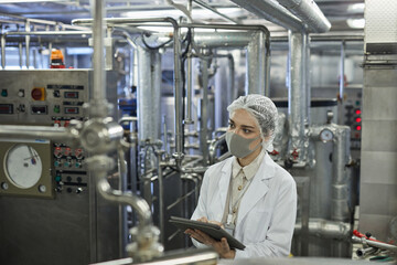 High angle portrait of young woman wearing mask and holding digital tablet during quality control inspection at food factory, copy space
