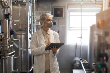 Waist up portrait of young woman wearing mask and holding digital tablet during quality control...