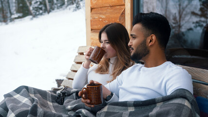 Close up. Happy young couple hugging covered with blanket on the outdoors bench. Drinking hot beverage and enjoying snow. High quality photo