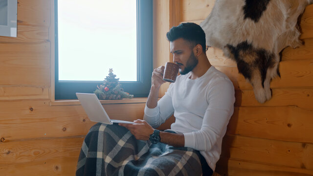  Young indian man sitting near the window in cozy house and working on laptop while drinking hot beverage. High quality photo