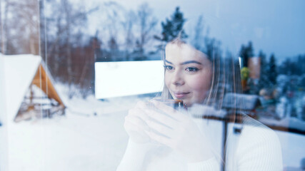 Young caucasian woman drinking hot beverage and looking through the window at snow covered houses....