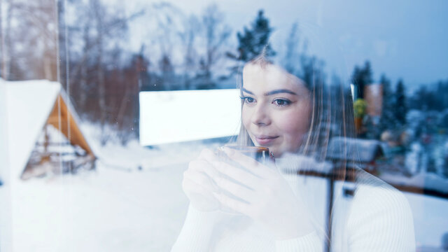 Young Caucasian Woman Drinking Hot Beverage And Looking Through The Window At Snow Covered Houses. High Quality Photo