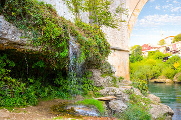 A small waterfall next to the historic Old Mostar Bridge or Stari Most with the village of Mostar, Bosnia and Herzegovina in view over the Neretva River.