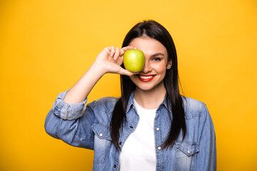 Happy caucasian woman leads a healthy lifestyle. A girl stands on an isolated orange background, holds a fresh green apple, looks into the camera covers one eye with an apple and smiles. Healthy food