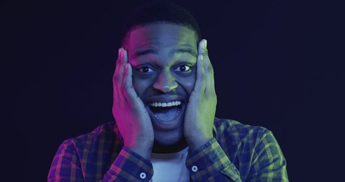 African american man feeling surprised and amazed, touching face in excitement and laughing, neon lights background