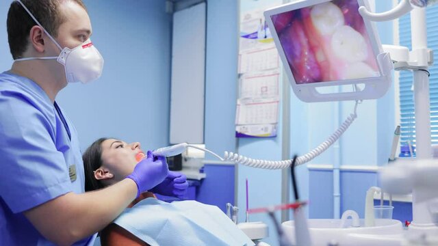 Male dentist is using a special camera which shows teeth image on a monitor for checking teeth health of female patient lying on dental chair. Visiting dentist, dental clinic