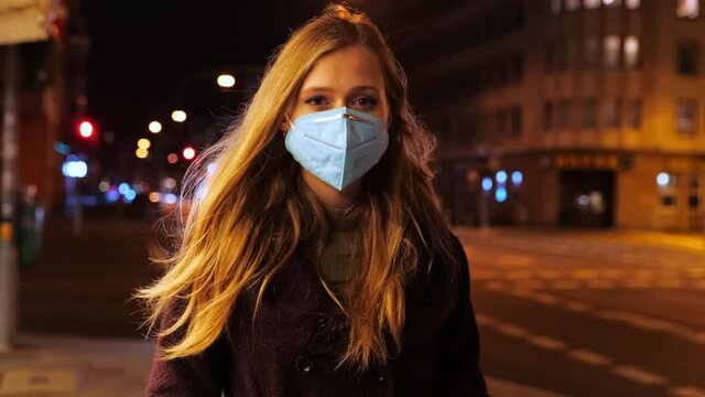 [4k] young woman wearing ffp2 face mask walking slowly at night on big city street in streetlight