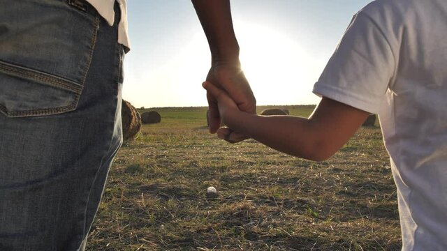 Close-up of joined human hands lit by setting sun, dark-skinned dad walking holding kid's hand. Big father's hand gently holding small child's hand during walk in countryside at sunset