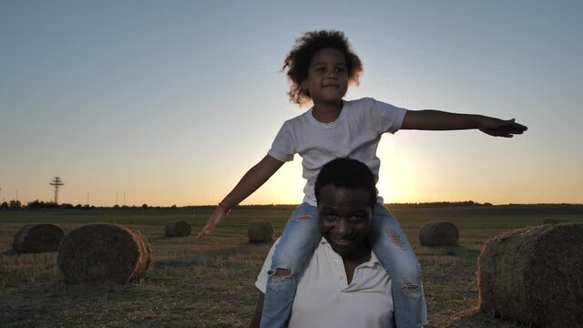 Cheerful african american man carrying happy, playing airplane, daughter while walking across harvested wheat field at sunset. Dark-skinned dad enjoying walk with preschool girl on his shoulders