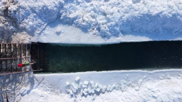 Winter swimming in Russia. View from above. A girl in a red cap with earflaps climbs out of the ice hole and climbs to the shore along the metal steps.