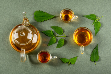 Cups of herbal tea, transparent teapot and nettle leaves on green background. Top view Flat lay