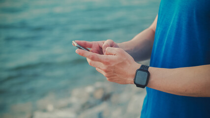 Close up hands typing a message on a smartphone. Man sports runner standing on the beach during the morning sunrise. Healthy lifestyle conception.