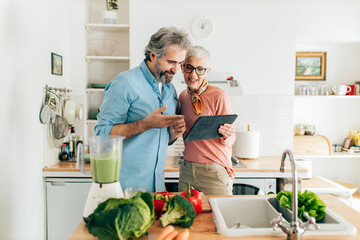 Senior couple preparing healthy smoothie in kitchen and using tablet to read recipe - 411684716