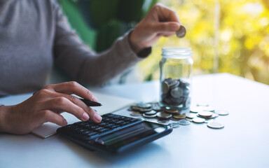 Closeup image of a woman putting coins in a glass jar and calculating on calculator for saving money and financial concept