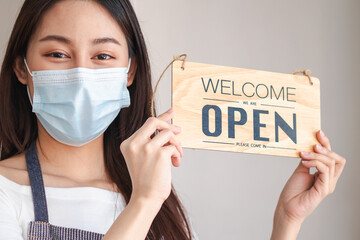 Smiling young asian owner retail,coffee shop woman holding sign board to open wearing face...