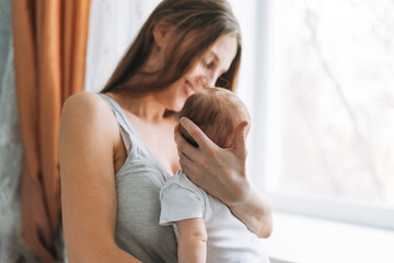 Young woman mom with long hair holding baby girl on hands near window at the home