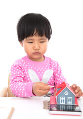 In front of the white background, the little girl is playing with the coins on the table and the model of the small house