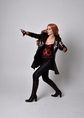 full length portrait of girl with long red hair wearing dark leather coat, corset and boots. Standing pose facing front on with  magical hand gestures against a  studio background.
