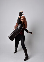 full length portrait of girl with long red hair wearing dark leather coat, corset and boots. Standing pose facing front on with  magical hand gestures against a  studio background.