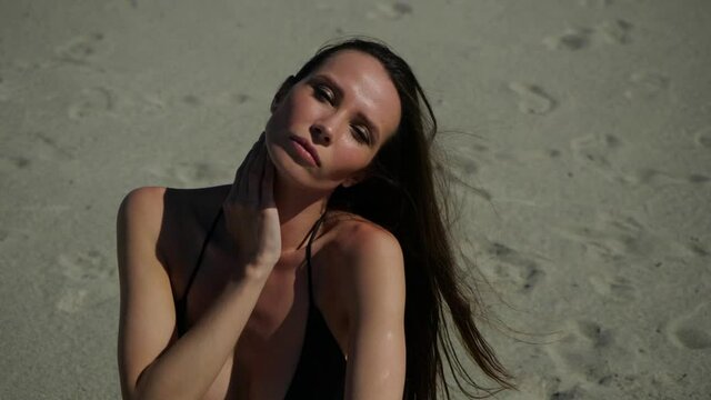 Sexy young girl in a black bikini sits on the sand staring into the camera and sensually strokes her body on a warm summer day. High quality FullHD footage