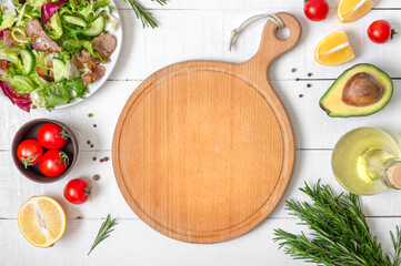 Mockup with empty wood cutting board. Fresh salad and ingredients for cooking on white wooden...