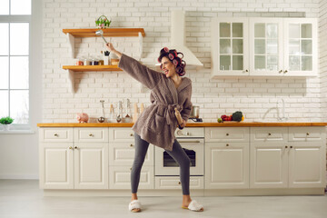 Happy housewife having fun while making breakfast. Beautiful young woman in hair curlers, bathrobe, leggings and house slippers holding cooking utensils, dancing and singing songs in spacious kitchen