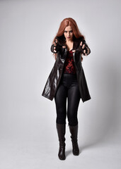 full length portrait of girl with long red hair wearing dark leather coat, corset and boots. Standing pose facing front on with  magical hand gestures against a  studio background, low camera angle.