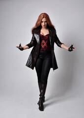 full length portrait of girl with long red hair wearing dark leather coat, corset and boots. Standing pose facing front on with  magical hand gestures against a  studio background, low camera angle. © faestock