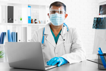 Middle-aged male doctor in face shield and mask sitting at the table at his office