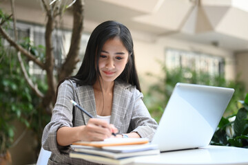 Businesswoman working with laptop computer and writing something in her schedule book at outdoor workplace.