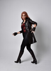 full length portrait of girl with long red hair wearing dark leather coat, corset and boots. Standing pose facing front on with  magical hand gestures against a  studio background, low camera angle.