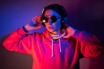 cyberpunk woman in a hooded hoodie and sunglasses dances against a wall with neon sticks hanging around her neck