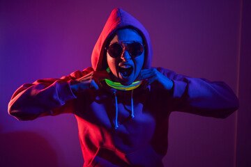 cyberpunk woman in a hooded hoodie and sunglasses dances against a wall with neon sticks hanging around her neck