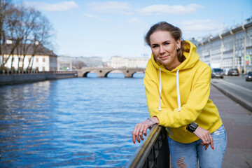 Cute young woman stands on the embankment in a yellow sweatshirt