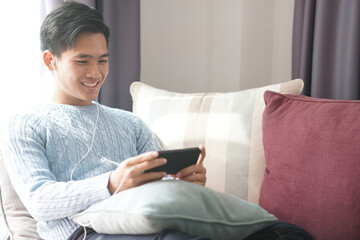 asian man wearing earphones listening to music on smart phone relaxing on sofa at home