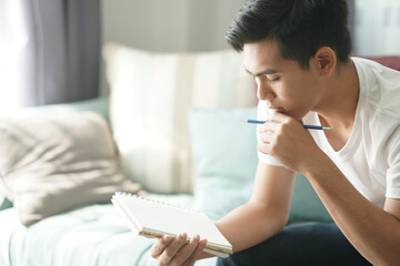 thoughtful asian young student man teenager thinking holding notebook on sofa at home