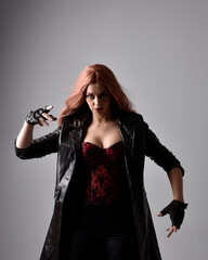 Close up portrait of girl with long red hair wearing dark leather coat, corset and gloves. Posing with gestural hand movements as if casting spell. Moody back lit lighting against a studio background.