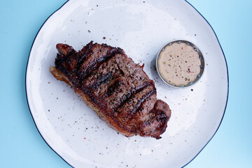 Delicious beef steak with sauce served on a white plate over pastel blue background. BBQ meat, barbecue concept.