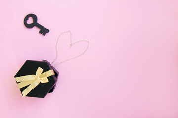 One dark black gift box with luxury gold bow with chain lined in the shape of a heart and a black wooden key on pink background. Horizontal with copy space. Flat lay