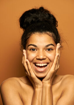 Look cute. Portrait of surprised young mixed race woman with perfect skin and hair in a bun looking excited, touching her face, posing isolated over orange background. Beauty, skincare concept