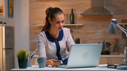 Hispanic woman shopping online late at night sitting in the kitchen on laptop using credit card. Employee making paymant transaction from home on digital notebook conected to internet
