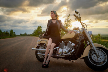 Fototapeta premium Attractive female brunette motorcyclist with motorcycle in a summer evening during sunset. Beautiful girl on nature and sky with sun and clouds background. Adventure concept