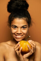 Portrait of excited naked young mixed race woman looking at camera, biting her lip, holding little orange pumpkin, posing isolated over orange background. Healthy eating, skincare concept