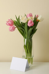 Vase with tulips and blank card against beige background, space for text