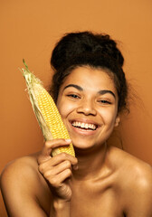 For healthy body. Portrait of happy naked young mixed race woman smiling at camera, holding yellow corn cob, posing over orange background. Healthy eating, vegetarian concept
