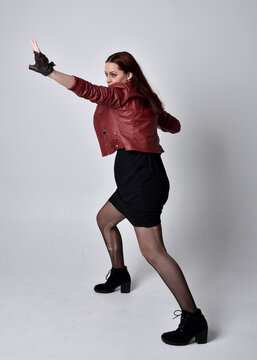 Full length portrait of girl wearing red leather jacket, tights with boots and gloves. Posing with gestural hand movements as if casting spell against a studio background.