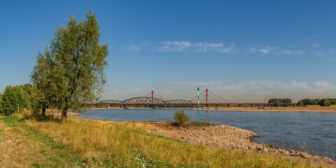 Obraz premium Duisburg, North Rhine-Westfalia, Germany - August 07, 2018: A parched meadow at the River Rhine with the Beeckerwerther Bridge and the Haus-Knipp Railway bridge in the background