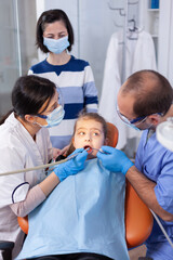 Child with mouth open looking at dentist assistant in the course of cavity treatment. Mother with her kid in stomatology clinic for teeth examine using modern instruments.